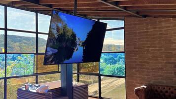 Una habitación moderna con una cama grande, un televisor en la pared y vistas a un paisaje verde a través de ventanas amplias.
