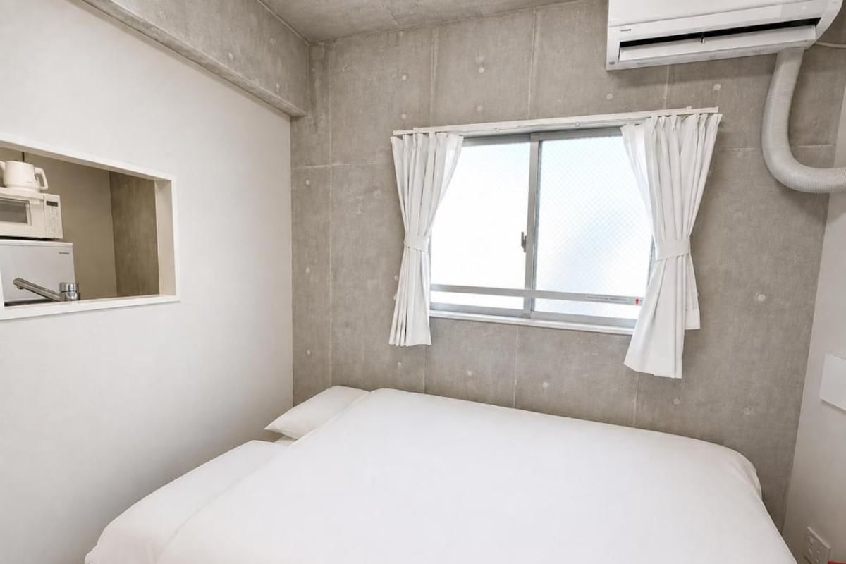 Un dormitorio con cama, ventana con cortinas, estante empotrado con microondas y unidad de aire acondicionado.