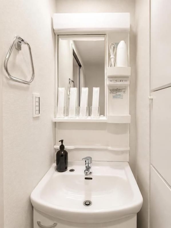 A bathroom with a white sink, a mirror, a towel rack, and a shelf with toiletries. - SLEEPLAB Ikebukuro (Toshima City)