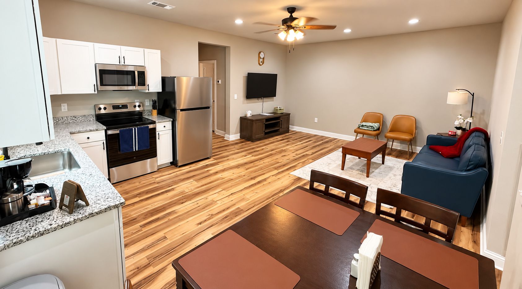 A modern kitchen with stainless steel appliances, a dining area with wooden tables and chairs, and a living area with a blue sofa and a ceiling fan.