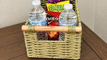 A wicker basket containing two bottles of water, a bag of FunJunk chips, and a bag of Doritos, with a remote control on a wooden table.