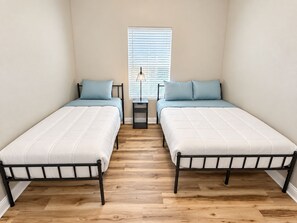 A bedroom with two single beds, a nightstand with a lamp, and a window with blinds. - Cozy 2-bedroom house in perfect Lufkin with AC for relaxing stays (Lufkin)