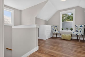 A room with a sloped ceiling, a white dresser, two floral chairs, a small ottoman, and a window with a view of trees. - Boise 1BR w/ Patio Near BSU, Downtown & Airport (Boise)