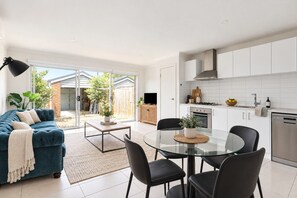 A modern living room with a sofa, a glass dining table, and chairs. There is a kitchen area with white cabinets and appliances. - Urban Pause on Flourish (Doreen)
