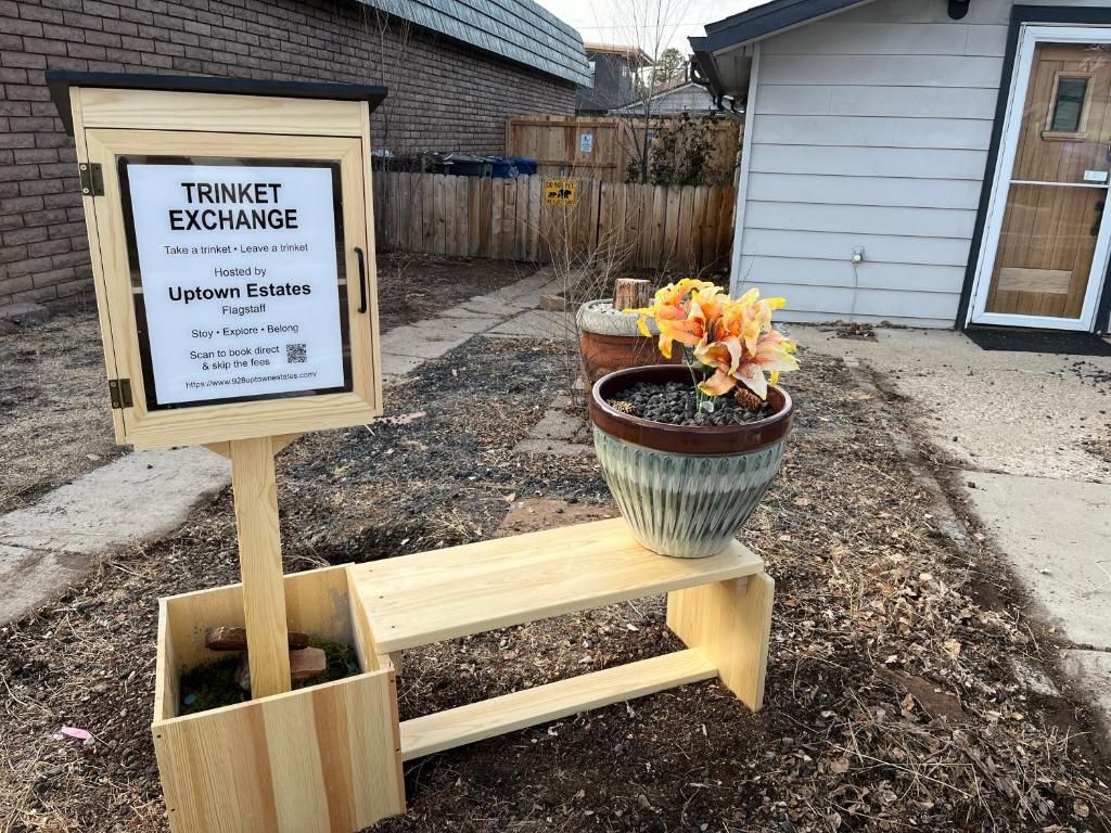 A wooden signboard with text for a trinket exchange, a planter with flowers, and a wooden bench.