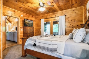 Ein Schlafzimmer einer Holzhütte mit einem Bett, einem Schminktisch, einem Deckenventilator und einem Fenster mit Vorhängen.