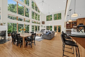 Een ruime woonkamer met een eettafel, stoelen en een bank. Grote ramen zorgen voor daglicht en uitzicht op het omliggende landschap.