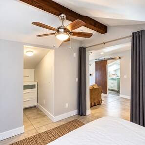 Quarto com ventoinha no teto, uma cama, mesa de cabeceira e vista para o banheiro adjacente.