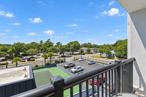 Blick vom Balkon auf einen Parkplatz mit mehreren Autos, einen Tennisplatz und umliegende Grünflächen.