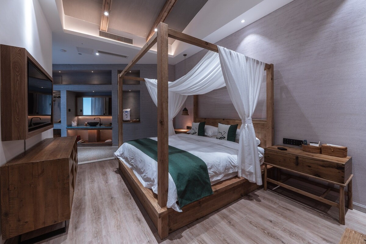 A modern bedroom with a four-poster bed, wooden furniture, and a view to the living area.
