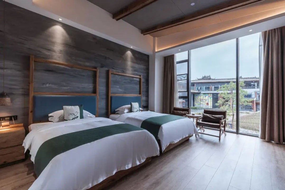 A hotel room with two beds, a small table, and a view of the outside.