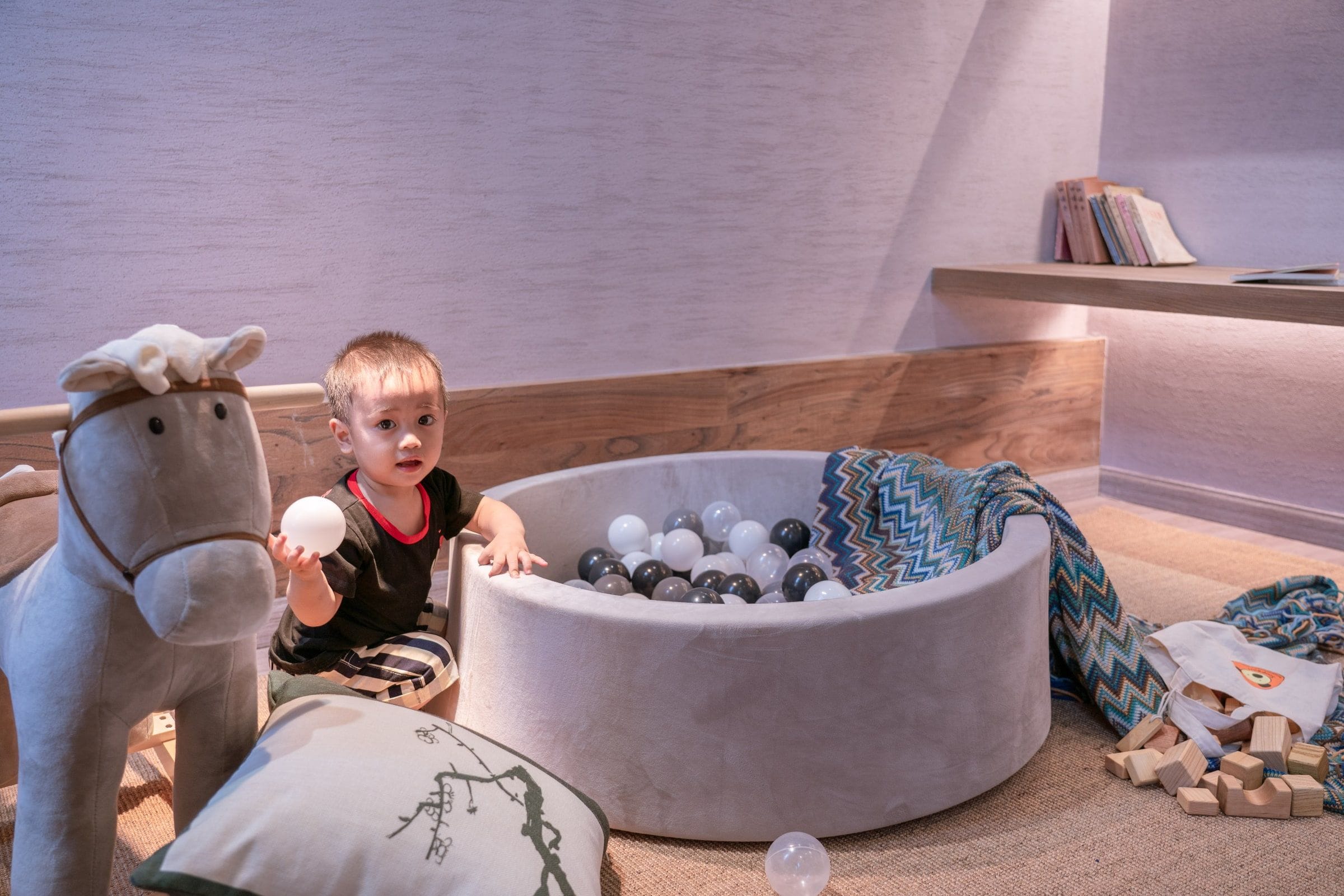 Niño jugando con una pelota en una habitación con un área de estar circular y acolchada llena de pelotas, un peluche grande y una manta estampada.