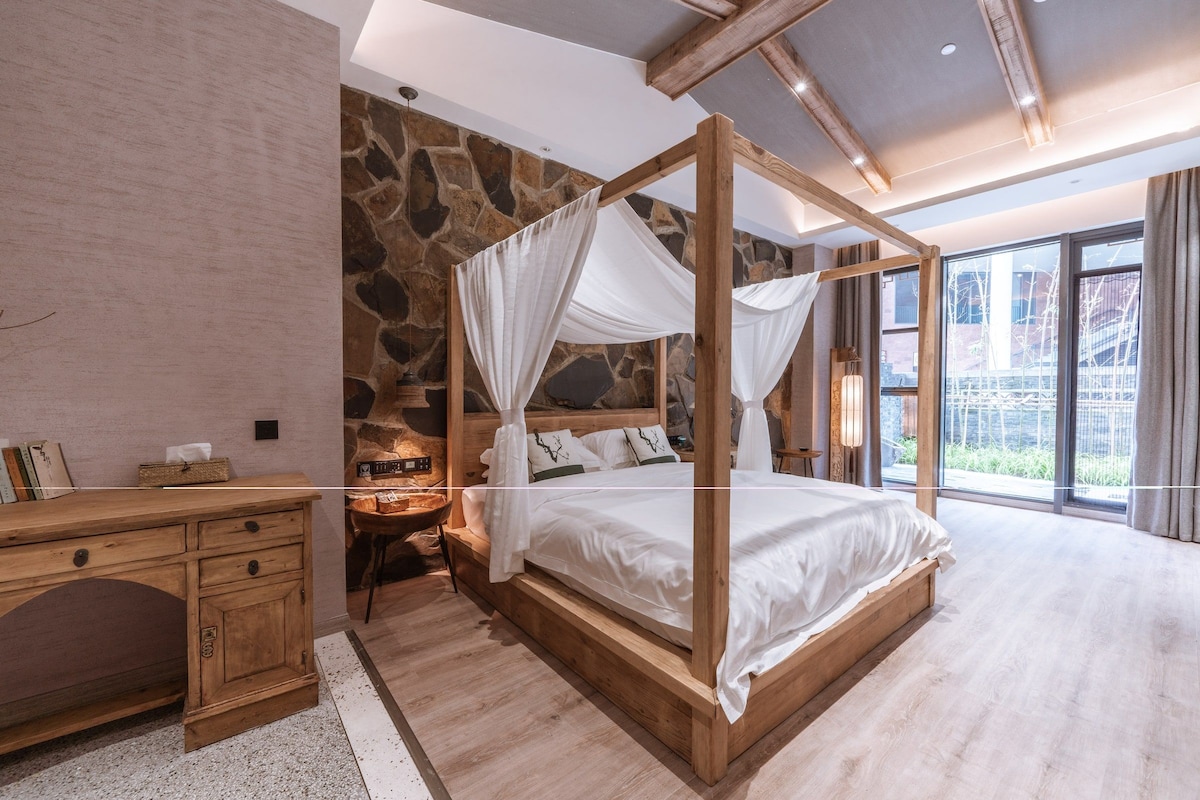 A bedroom with a four-poster bed, a wooden desk, a stone wall, and a large window.