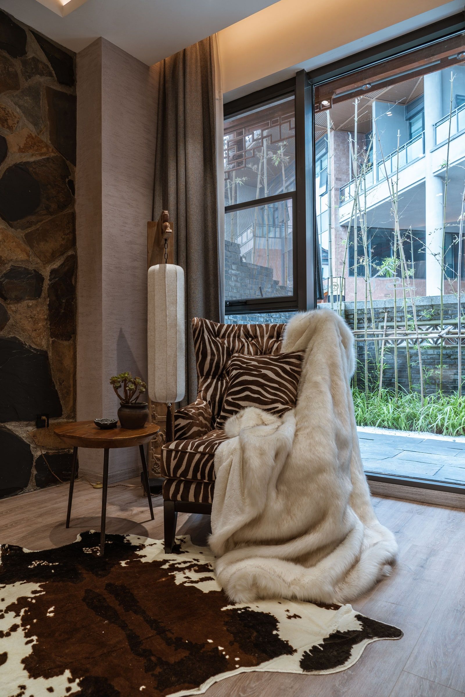 A cozy room with a stone wall, a zebra-patterned chair, a large window with a view of bamboo outside, and a cowhide rug on the floor.