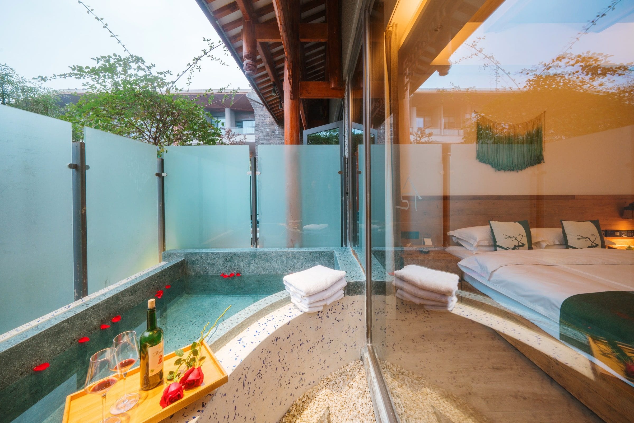 Un dormitorio moderno con una cama grande, un baño con paredes de vidrio y una bañera de hidromasaje, y un balcón con una mesa de madera y toallas.