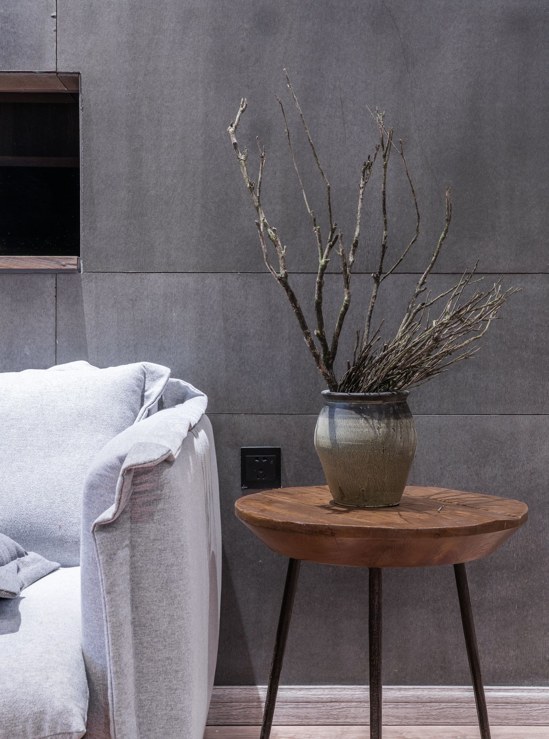 Una habitación moderna con un sofá gris, una mesa redonda de madera con un jarrón de ramas y una pared gris.