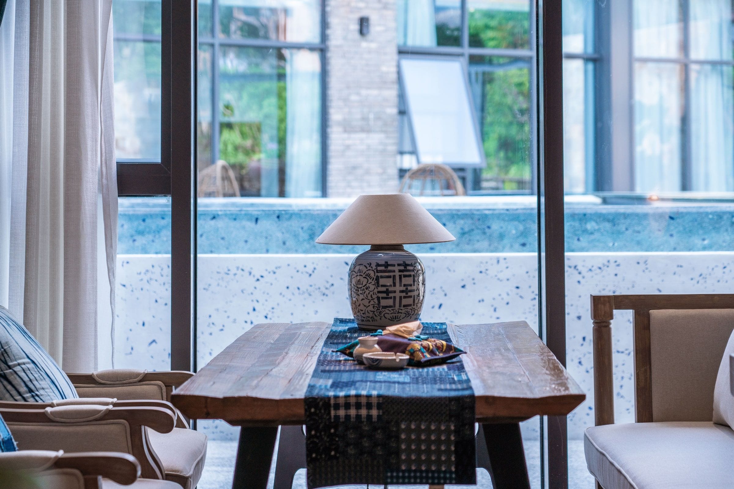 Una mesa con una lámpara, un posavasos y un cuenco de fruta en una habitación con vistas a una piscina y follaje exterior.