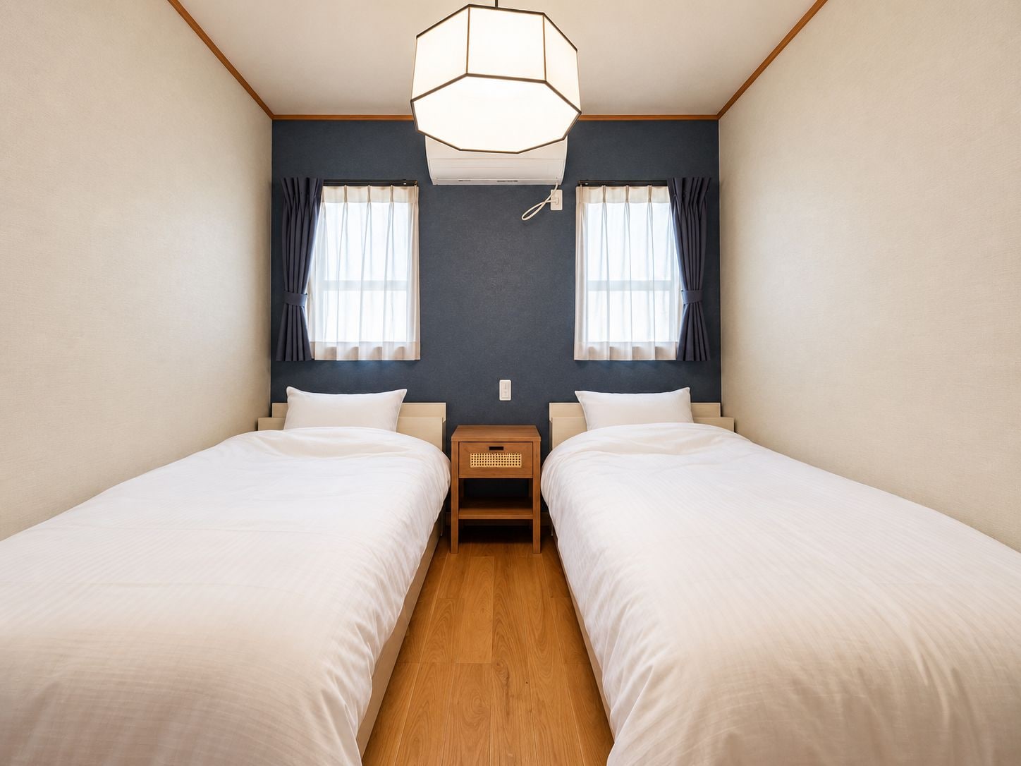 A hotel room with two beds, a wooden floor, a ceiling light fixture, and a nightstand between the beds.