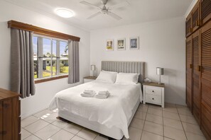 A bedroom with a bed, two bedside tables, a ceiling fan, a window with curtains, and a view of the outside. - Peaceful 2-Bedroom in Innisfail Cassowary Coast (Belvedere)
