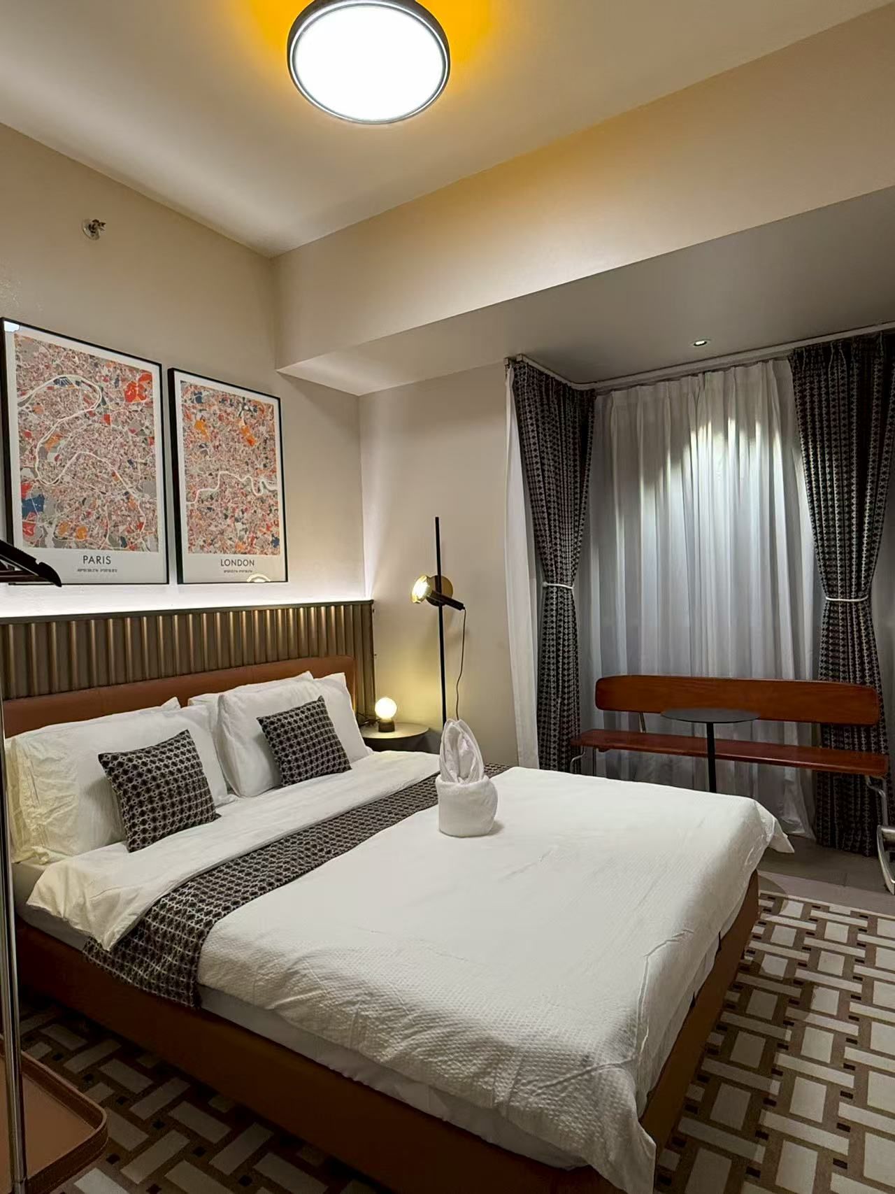 Una habitación de hotel con una cama, una mesita de noche, una lámpara, un banco y dos mapas enmarcados de París y Londres.