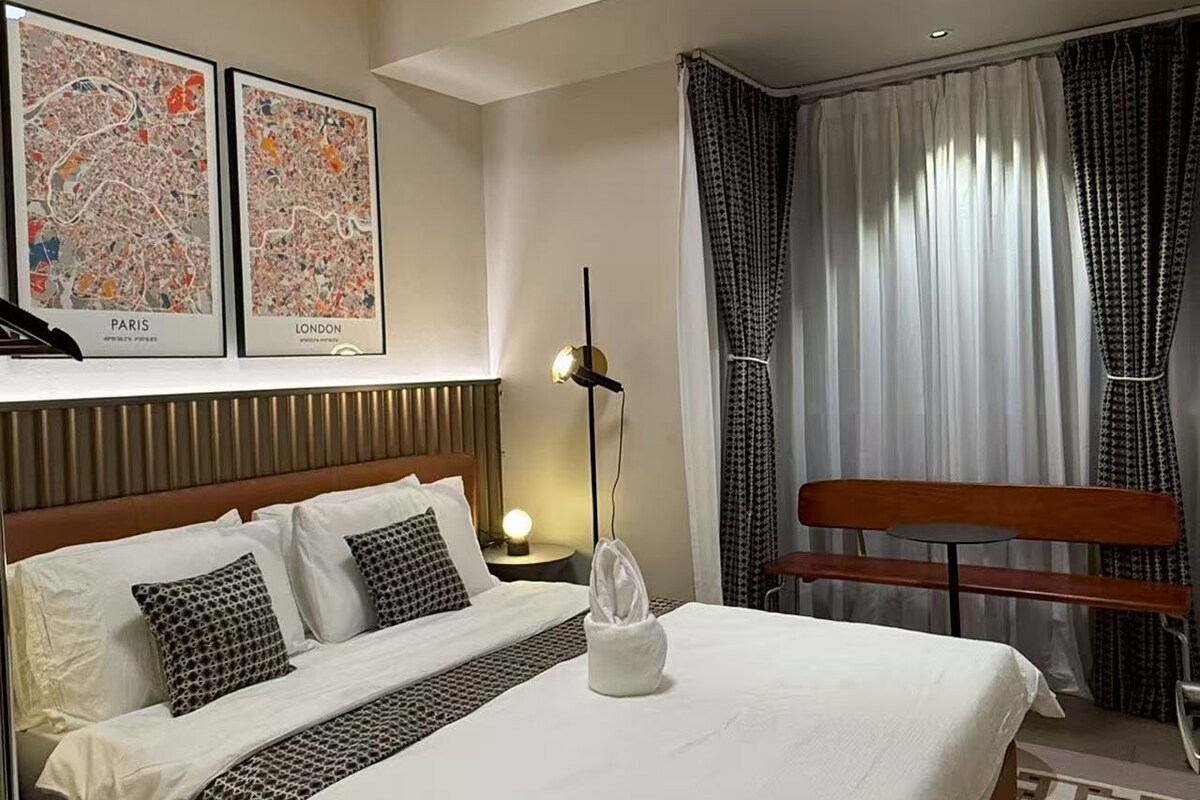 A hotel room with a bed, a nightstand, a lamp, a bench, and two framed maps of Paris and London.