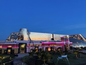 Un hotel moderno con una fachada de vidrio curva distintiva, un jardín cuidado y una entrada destacada con un dosel iluminado en rosa.
