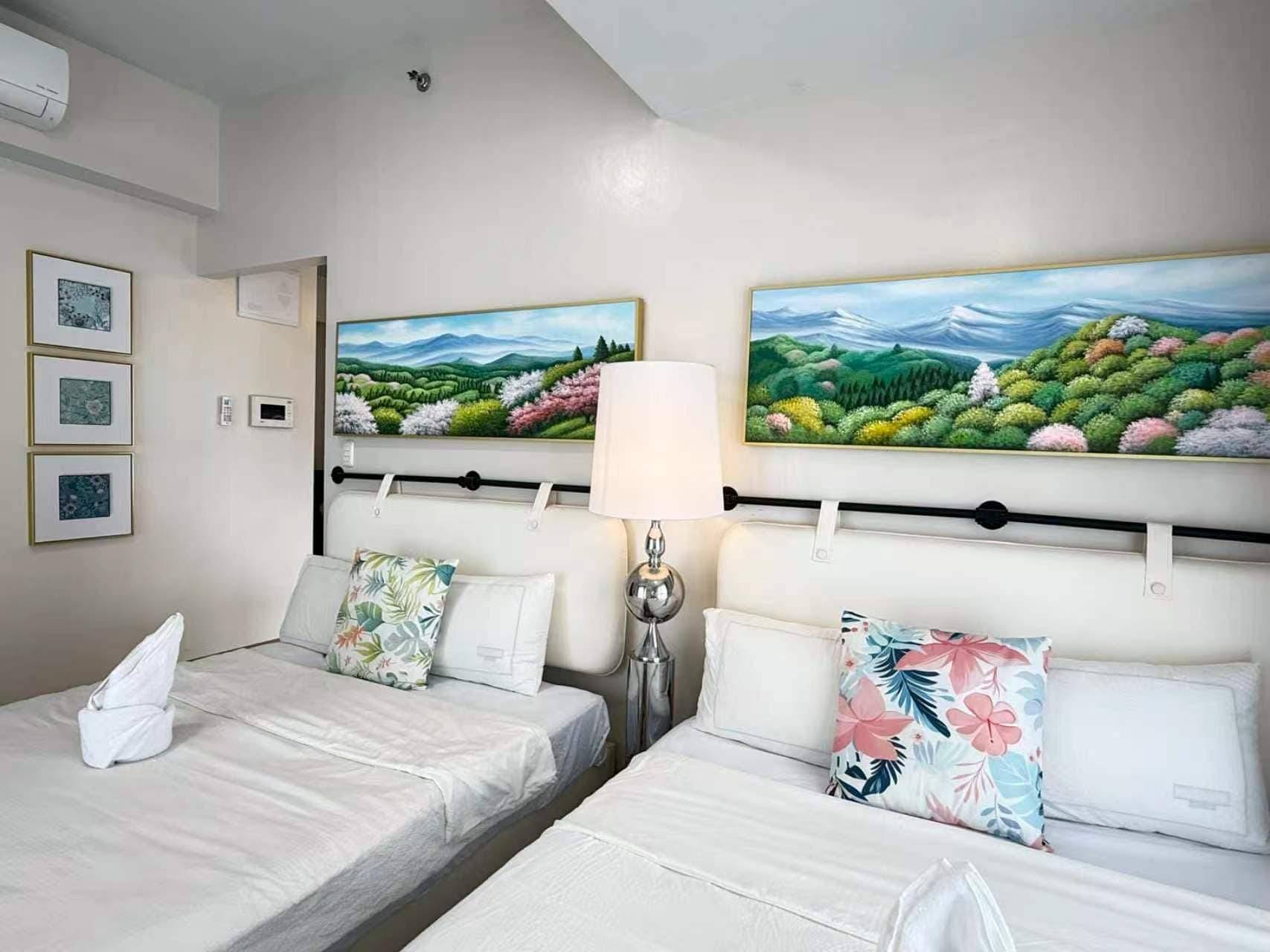 Une chambre d’hôtel avec deux lits, une table de chevet, une lampe et deux tableaux accrochés au mur.