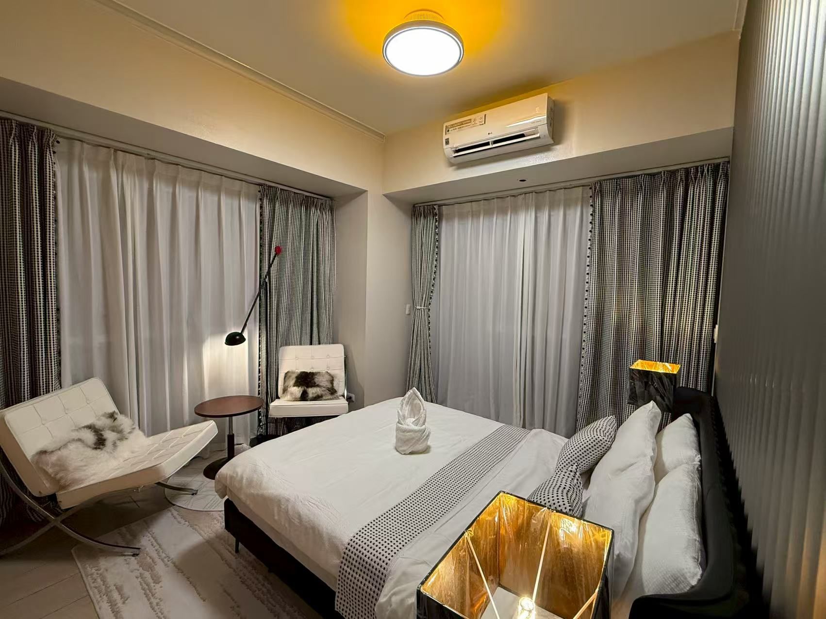 A hotel room with a bed, a chair, a nightstand, an air conditioning unit, and a lamp.