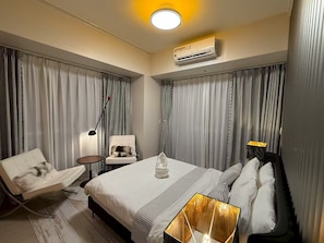 A hotel room with a bed, a chair, a nightstand, an air conditioning unit, and a lamp. - Seaview 1BR  2BR 3BR Next Okada (Parañaque)