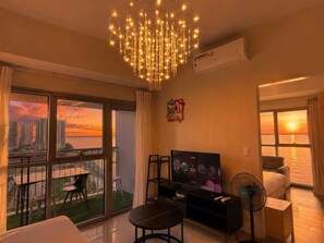 Habitación de hotel con balcón, televisión, ventilador y vista al atardecer sobre el agua.