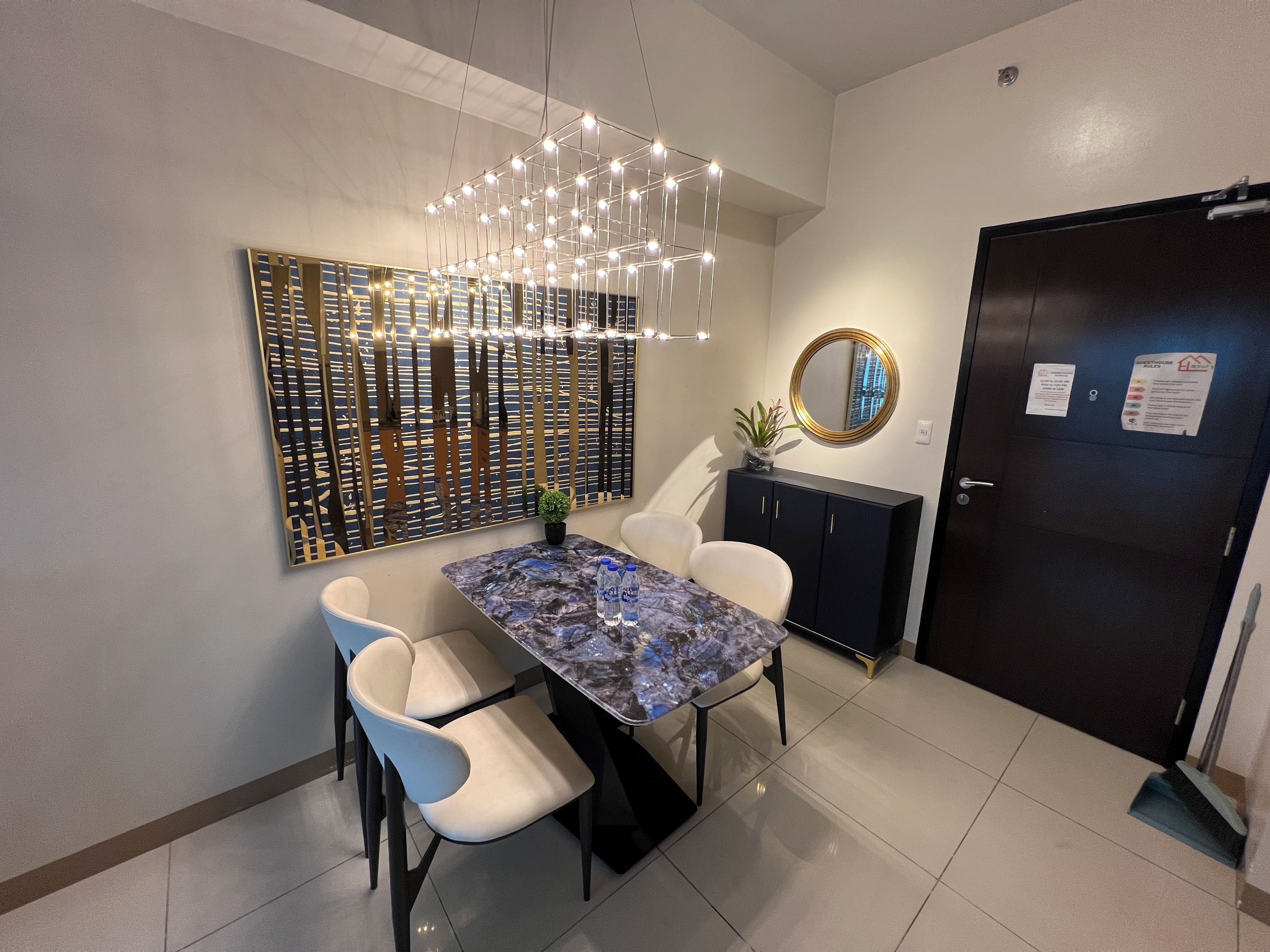 Un comedor con una mesa de mármol, sillas blancas, un accesorio de iluminación decorativo, un armario negro y una puerta con un cartel.
