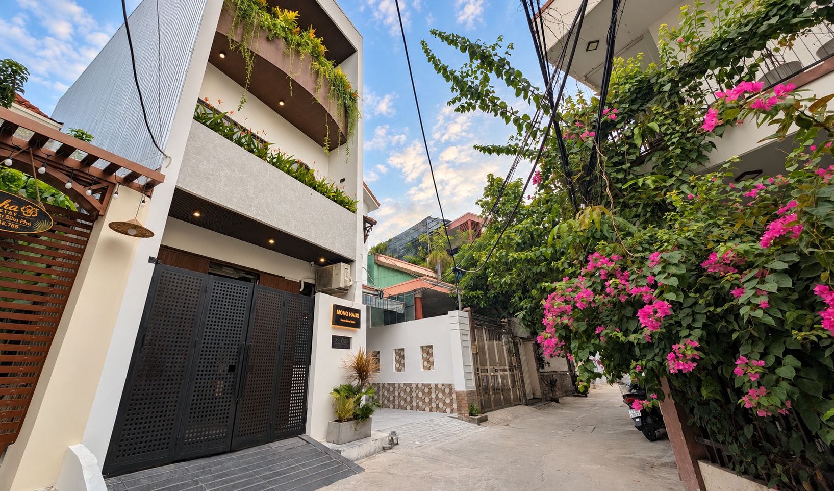 A modern two-story building with a gated entrance, a sign indicating 'MONO HAUS,' and a lush garden with flowering plants.
