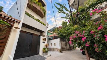 A modern two-story building with a gated entrance, a sign indicating 'MONO HAUS,' and a lush garden with flowering plants.