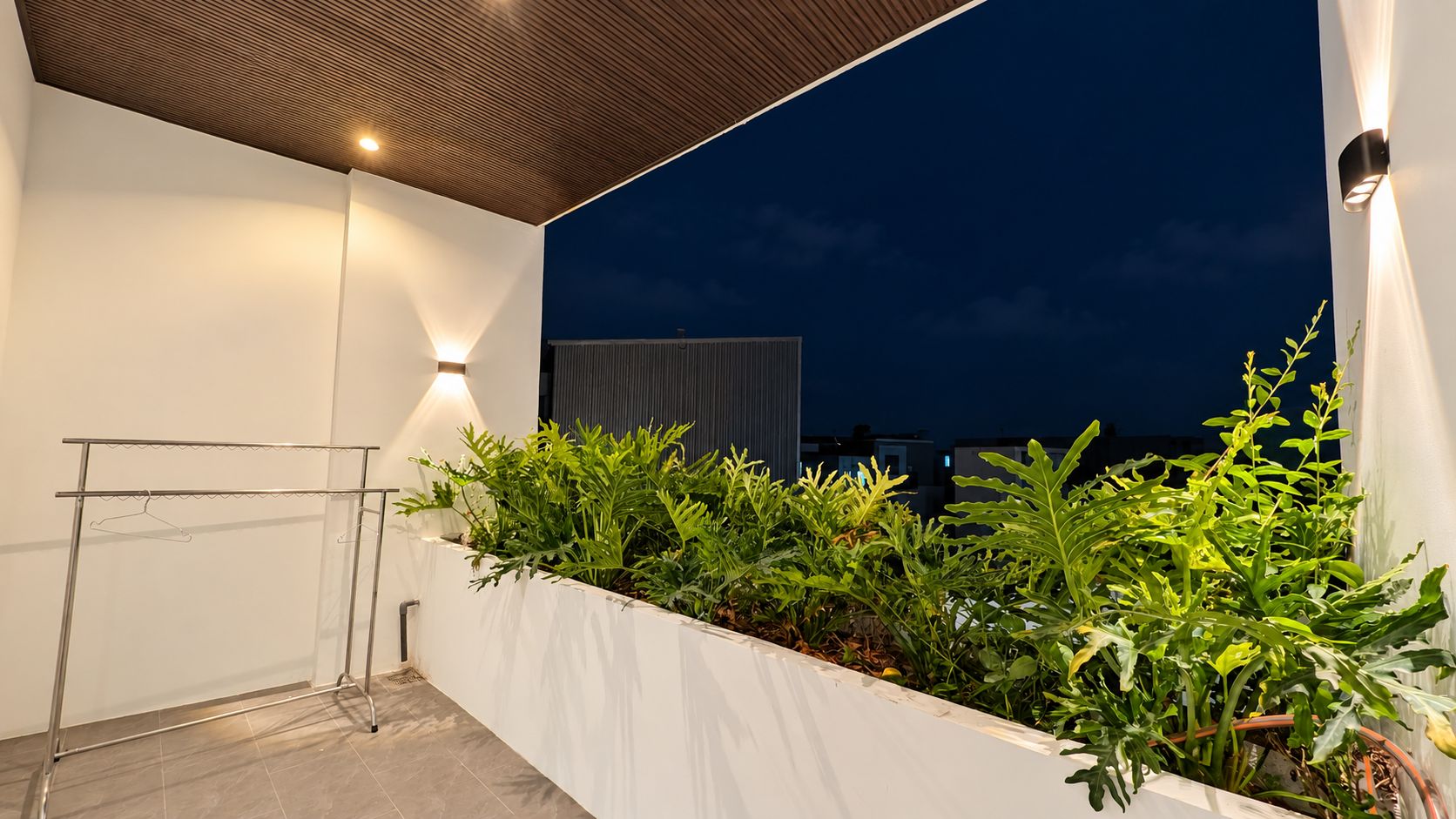 An outdoor balcony with a metal drying rack, a white planter with green plants, and a night sky.
