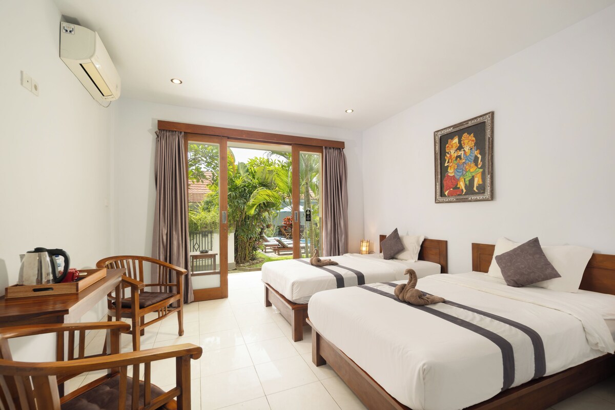 A hotel room with two beds, a desk with a kettle, a chair, and a painting on the wall.