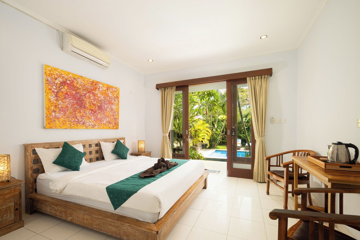 A hotel room with a wooden bed, a painting, a view of a swimming pool, and a dining area with a table and chairs.