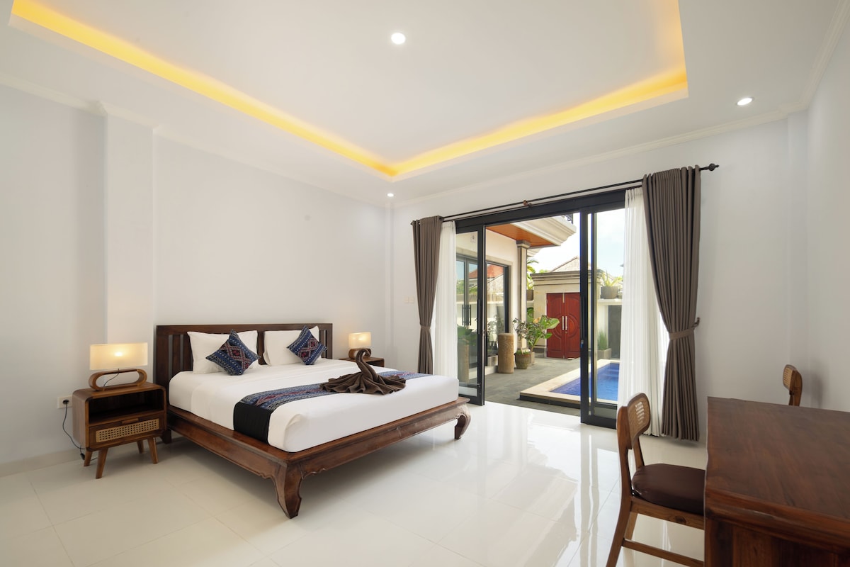 A hotel room with a large bed, a wooden nightstand, a table, and a view of a swimming pool through a glass door.