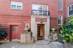 A red brick building with a central entrance, a red door, and a balcony with a wrought iron railing.