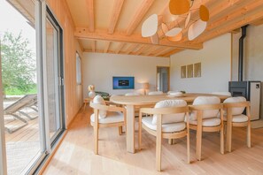 A spacious dining area with a large wooden table and white cushioned chairs, a flat-screen TV mounted on the wall, and a wood-burning stove. - Au Bout du Chemin Ecolodge (GERAUDOT)