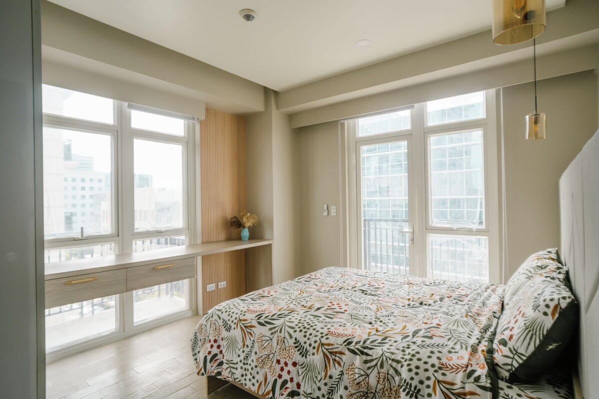 Ein Schlafzimmer mit einem großen Fenster, einem Bett mit einer blumigen Tagesdecke und einem Schreibtisch mit einer Vase.