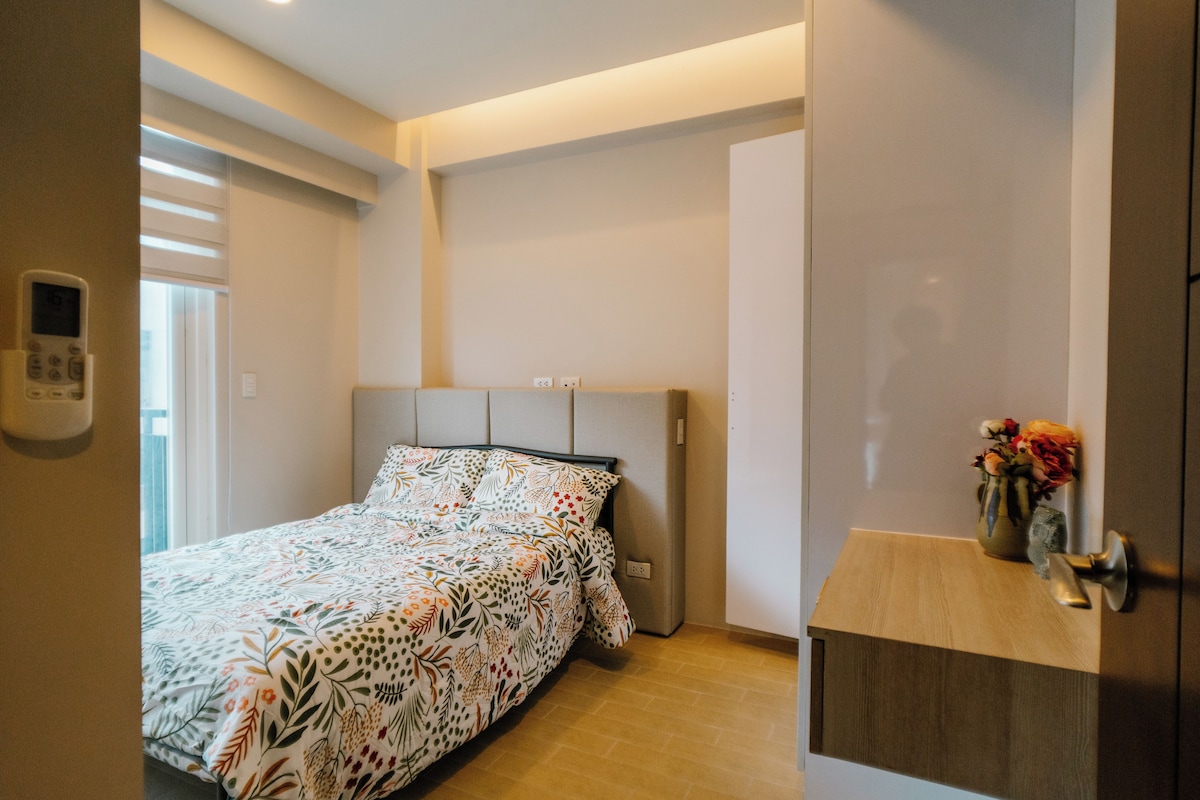 Ein Schlafzimmer mit einem Bett, einem Nachttisch mit Blumenvase, einem an der Wand montierten Thermostat und einer Tür mit Griff.
