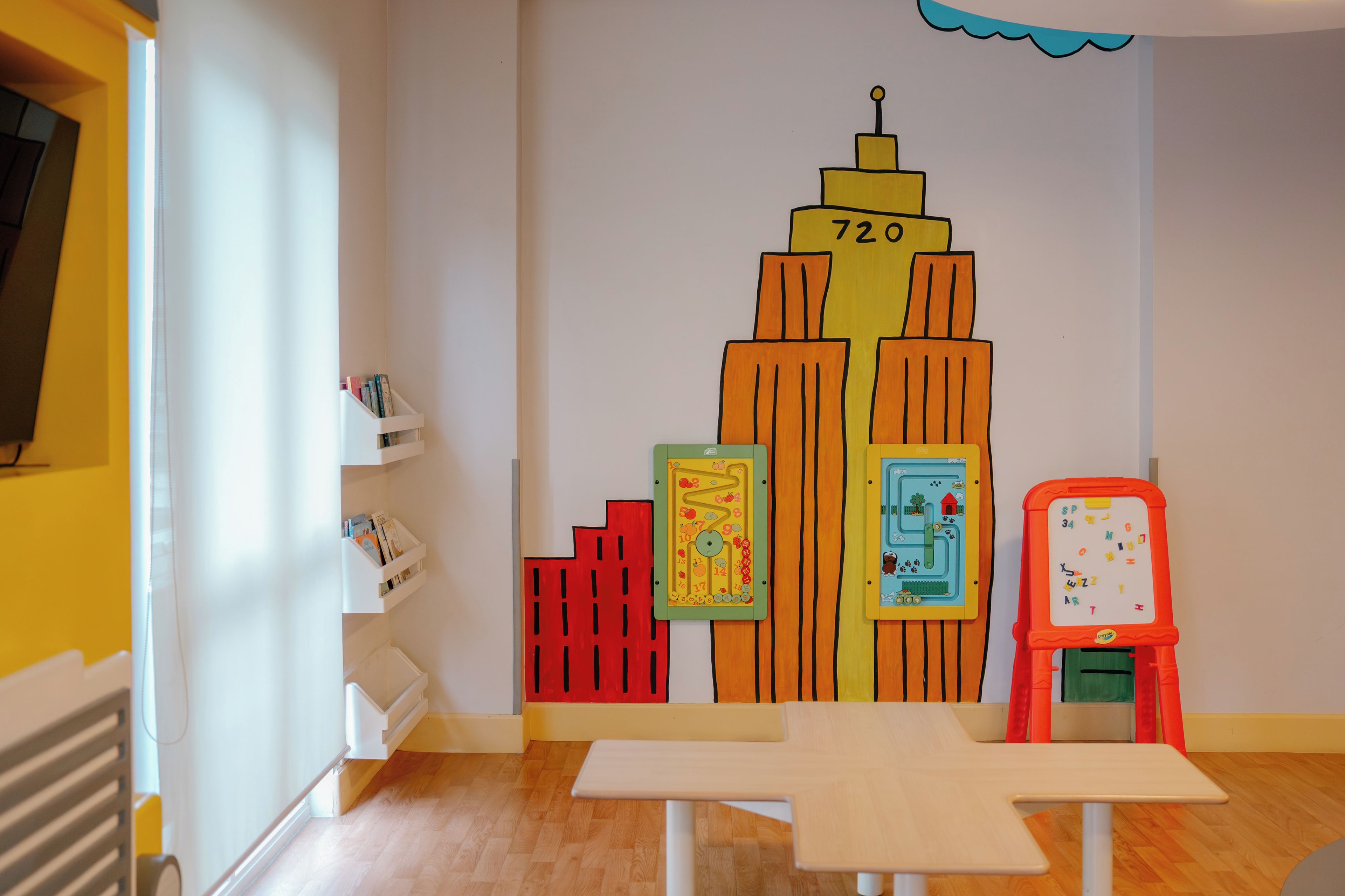 A children's play area with a colorful mural, a white shelf with books, and a small table.