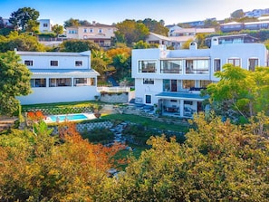 Una casa moderna de dos plantas con piscina, rodeada de abundante vegetación y otras edificaciones residenciales.