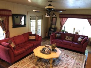 A living room with two red sofas, a coffee table with books and a plant, a ceiling fan, and a large window with curtains. - Rocky River | Near Town w/ Wildlife & Large Patio (Bandera)