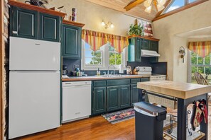 A kitchen with wooden cabinets, a dishwasher, a stove, and a window with curtains. - Felix Cabin at Restoration Ranch (Fredericksburg)