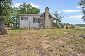 A single-story house with a stone chimney, a small porch, and a grassy yard. - Freida Cabin at Restoration Ranch (Fredericksburg)