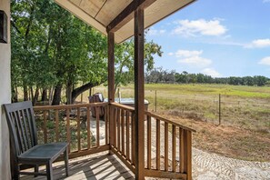 A wooden porch with a bench and table, overlooking a grassy field and trees. - Freida Cabin at Restoration Ranch (Fredericksburg)