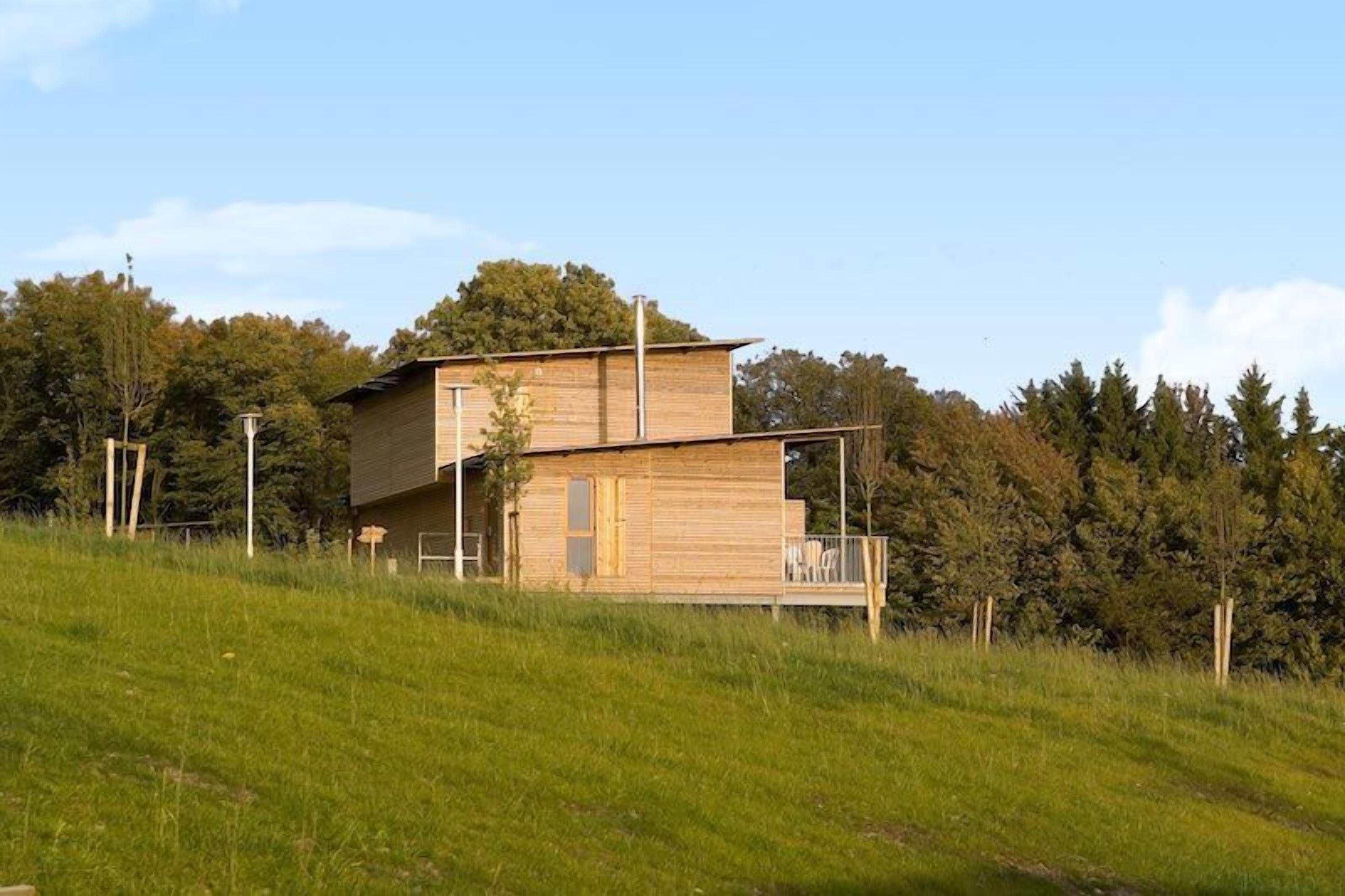 A modern two-story house with a flat roof and a wooden exterior, situated on a grassy hill with trees in the background.