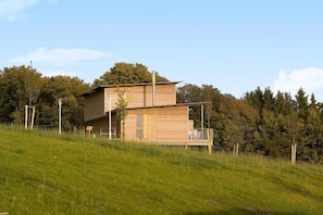 A modern two-story house with a flat roof and a wooden exterior, situated on a grassy hill with trees in the background. - Cottage en bois cosy | Terrasse privée! (Eppe Sauvage)