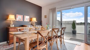 Uma área de refeições com mesa de madeira posta para quatro pessoas, acompanhada por duas luminárias, um violão e vista do céu através de grandes janelas.